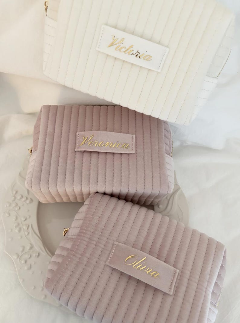 Puede incluir: Tres neceseres de pana en crema, malva y rosa empolvado. Cada bolsa tiene una etiqueta rectangular con un nombre en escritura dorada: "Victoria", "Veronica" y "Olivia". Las bolsas tienen cremallera y un dise&ntilde;o acolchado.