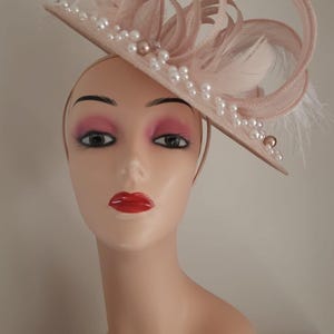 May include: A blush-colored hat with a flat brim, adorned with pearls, feathers, and looped fabric details. The hat is displayed on a mannequin head with red lipstick and eye makeup. A stylish accessory.