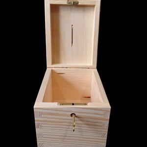 May include: A light-colored wooden box with a hinged lid and a vertical coin slot on the interior lid panel. The box features box joint corners, a metal hinge, and a small brass-colored key inserted into a circular keyhole on the front panel.