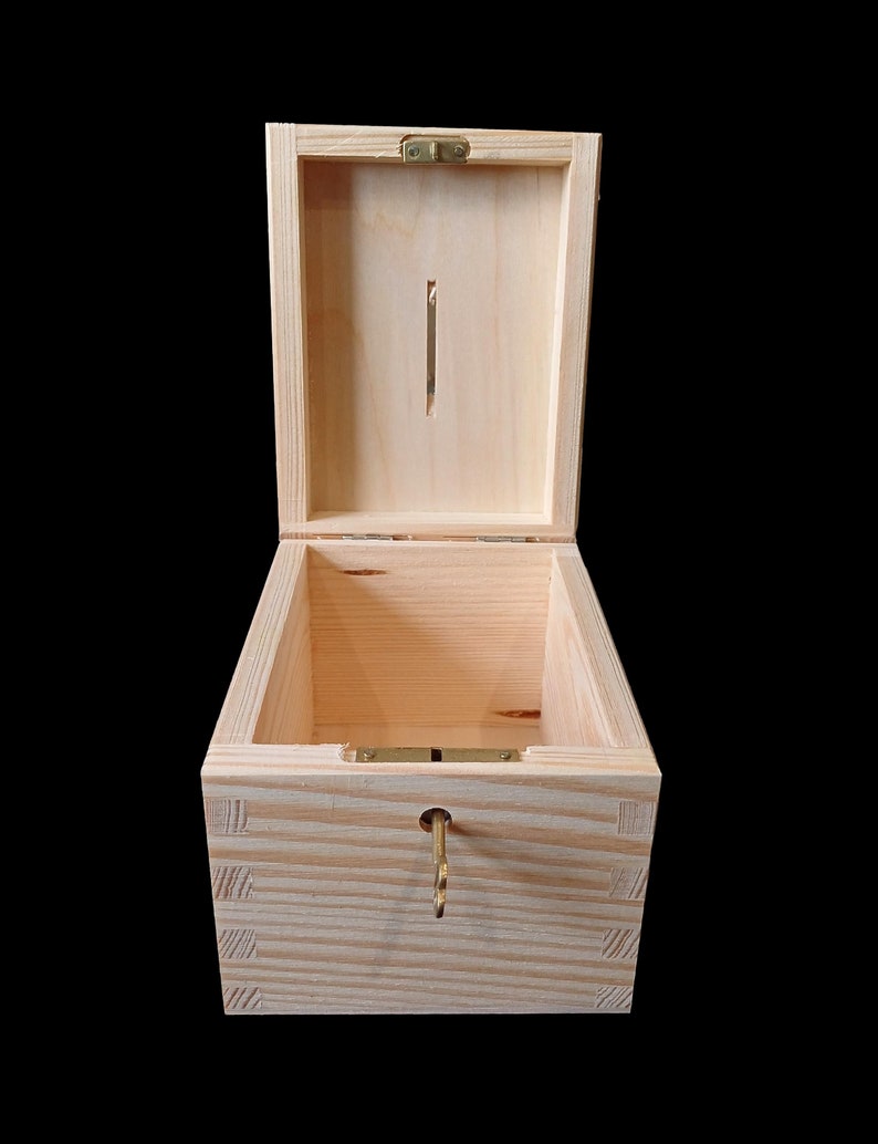 May include: A light-colored wooden box with a hinged lid and a vertical coin slot on the interior lid panel. The box features box joint corners, a metal hinge, and a small brass-colored key inserted into a circular keyhole on the front panel.