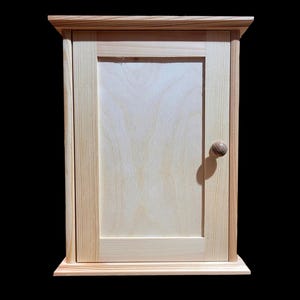 May include: A small, wooden cabinet with a single door and a round knob. The cabinet is made of light-coloured wood and has a simple, traditional design.