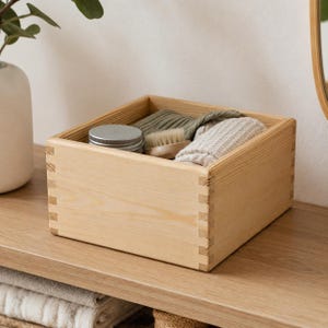 May include: A square light-coloured wooden storage box with visible finger joints at the corners. Inside the box, there is a small round metal container, a wooden-handled brush, and folded beige and olive green textured fabric items.