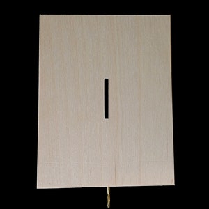 May include: A rectangular light-colored wooden box with a vertical slot centered on the front panel. A small metallic gold-colored latch or hook is visible protruding from the bottom edge of the wooden structure against a black background.
