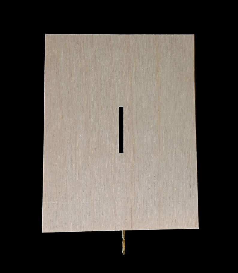 May include: A rectangular light-colored wooden box with a vertical slot centered on the front panel. A small metallic gold-colored latch or hook is visible protruding from the bottom edge of the wooden structure against a black background.