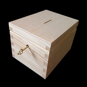 May include: A light-colored wooden box with a rectangular slot on the top lid. The box features visible finger joints at the corners and a small brass-colored key inserted into a circular keyhole on the front panel of the wooden container.
