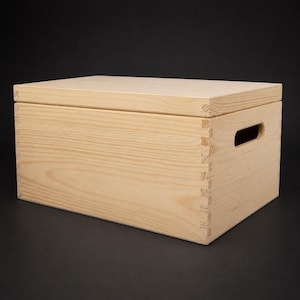 May include: A light-coloured wooden box with a lid, featuring finger-jointed corners and a handle cutout on one side. The box is made of unfinished wood, showcasing the natural grain and texture.