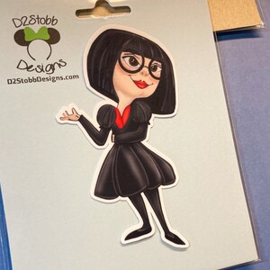 May include: A sticker featuring a cartoon character with black hair, wearing a black dress, red collar, and glasses. The character is standing with one arm raised and a smile on her face. The sticker is on a blue background with the text "D2Stobb Designs" and a website address.