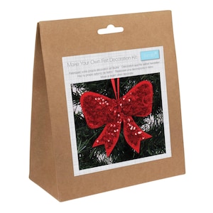 May include: A brown paper bag with a white label showing a red sequined bow decoration. The label reads "Make Your Own Felt Decoration Kit" in multiple languages. The bow is set against a backdrop of green foliage.