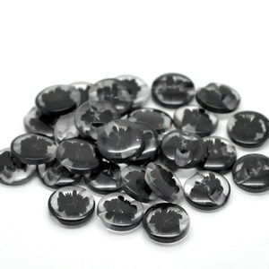 May include: A collection of clear, circular glass gems featuring a black floral pattern. The gems are scattered on a white background, displaying shades of grey and black. These gems are likely intended for decorative use.