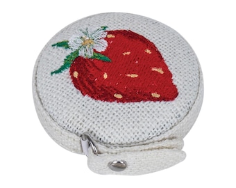 May include: A round, off-white tape measure with a red embroidered strawberry design. The strawberry has green leaves and a white flower. The tape measure has a silver metal zipper and a small fabric loop with a snap closure.