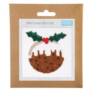 May include: A mini cross stitch kit featuring a Christmas pudding design. The pudding is brown with a white sauce, green holly leaves, and red berries. The kit is packaged in a brown cardboard backing.