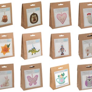 May include: A collection of craft kits in brown paper bags, each featuring a different felt ornament design. Designs include a fox, hedgehog, heart, chicken, flower, dinosaur, robot, sheep, bunny, butterfly, teapot, and owl. Each kit has the text "Make Your Own... Felt Ornament".