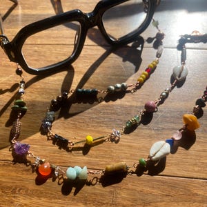 May include: A pair of black-framed eyeglasses attached to a long, decorative chain made of copper wire. The chain features a variety of colorful beads, including small cowrie shells, smooth round stones, faceted gems, and cylindrical metal spacers.