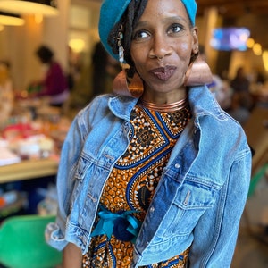 May include: A woman wearing a blue beret, a denim jacket, and a patterned dress with a teal bow. She has a nose ring and large copper earrings.