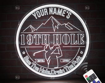 Puede incluir: Un letrero de neón blanco con el texto "YOUR NAME'S 19TH HOLE" y una silueta de golfista. El letrero también dice "ESTD. 1990 See The Hole, Drain The Putt".