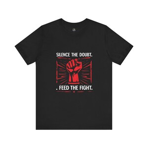 Puede incluir: Camiseta negra con un puño cerrado rojo dentro de un marco rectangular. El texto blanco "SILENCE THE DOUBT. FEED THE FIGHT." está impreso encima y debajo del gráfico.