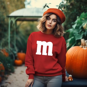 May include: A woman wearing a red sweatshirt with a white "M" on the front. She is standing in front of a pumpkin and wearing a red beanie.