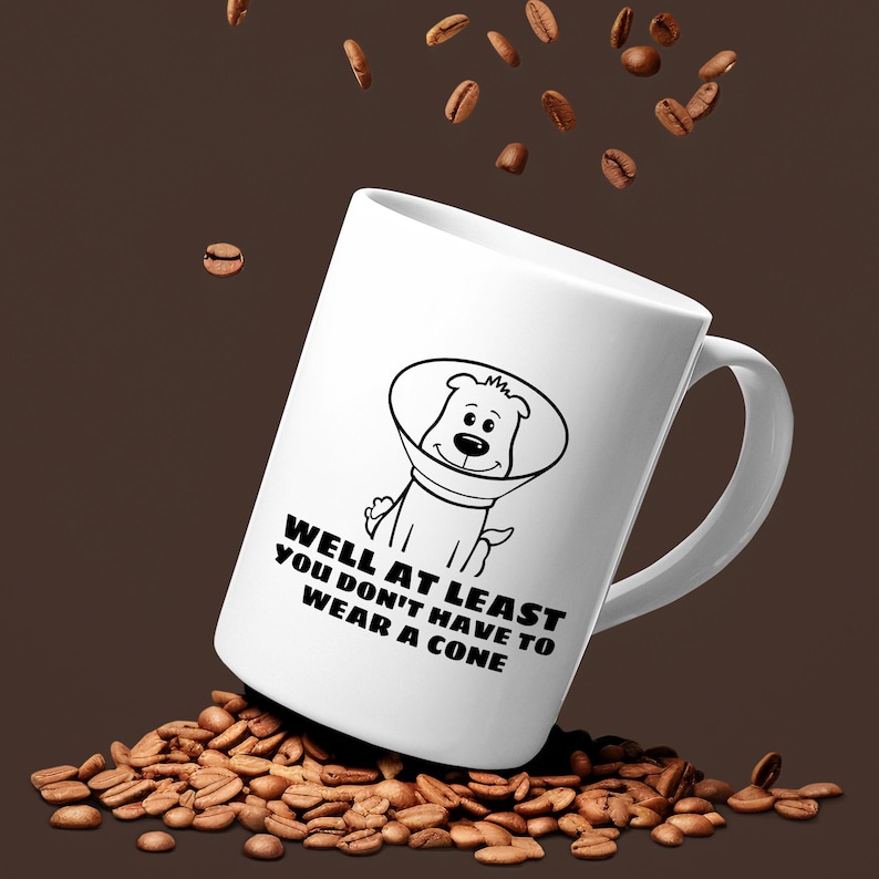 May include: A white ceramic mug featuring a black line drawing of a dog wearing a plastic cone collar. Below the illustration, the text reads: WELL AT LEAST YOU DON'T HAVE TO WEAR A CONE. The mug is surrounded by scattered roasted coffee beans.