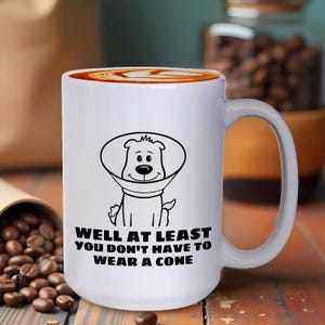 May include: A white ceramic mug featuring a black line drawing of a dog wearing a veterinary cone. Below the illustration, the text reads: WELL AT LEAST YOU DON'T HAVE TO WEAR A CONE. The mug has a smooth finish and a curved handle on the right side.