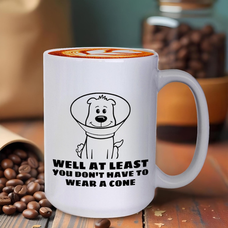 May include: A white ceramic mug featuring a black line drawing of a dog wearing a veterinary cone. Below the illustration, the text reads: WELL AT LEAST YOU DON'T HAVE TO WEAR A CONE. The mug has a smooth finish and a curved handle on the right side.