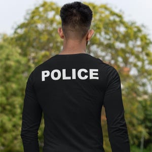 以下が含まれることがあります： 背中に大きな白い大文字で「POLICE」とプリントされた黒い長袖シャツ。人物はカメラから背を向けており、背景にはぼやけた緑の木々があります。