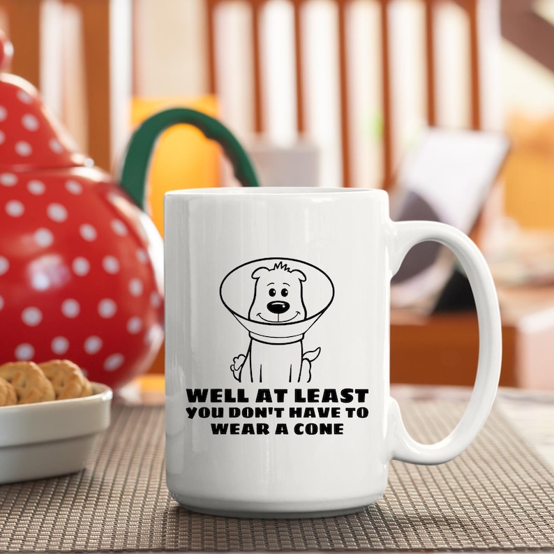 May include: A white ceramic mug featuring a black line drawing of a dog wearing a veterinary cone. Below the illustration, the text reads: WELL AT LEAST YOU DON'T HAVE TO WEAR A CONE. The mug has a standard handle and a smooth, cylindrical body.