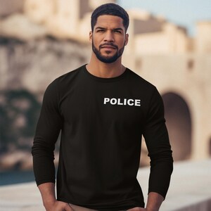 以下が含まれることがあります： 胸に白で「POLICE」とプリントされた黒の長袖シャツ。シャツは柔らかく不透明な素材で作られています。シャツはクルーネックです。