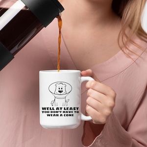 May include: A white ceramic mug featuring a black line drawing of a dog wearing a veterinary cone. Below the illustration, the text reads: WELL AT LEAST YOU DON'T HAVE TO WEAR A CONE. The mug has a curved handle and is being filled with a dark brown liquid.