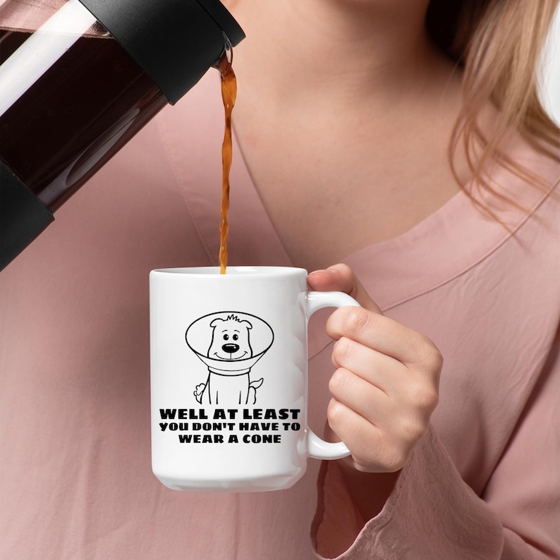 May include: A white ceramic mug featuring a black line drawing of a dog wearing a veterinary cone. Below the illustration, the text reads: WELL AT LEAST YOU DON'T HAVE TO WEAR A CONE. The mug has a curved handle and is being filled with a dark brown liquid.
