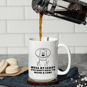 May include: A white ceramic mug with a black line drawing of a dog wearing a cone collar. Below the illustration, the text reads: WELL AT LEAST YOU DON'T HAVE TO WEAR A CONE. Dark coffee is being poured into the mug from a glass French press.