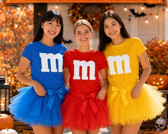 May include: Three short-sleeved t-shirts in blue, red, and yellow, each featuring a large white lowercase letter m on the chest. Each shirt is paired with a matching colored tulle skirt that includes a decorative bow at the waistline.