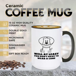 May include: A white 15 oz ceramic coffee mug with a black handle and rim. The mug features a black line drawing of a dog wearing a cone collar with the text: WELL AT LEAST YOU DON'T HAVE TO WEAR A CONE. The mug is microwave and dishwasher safe.