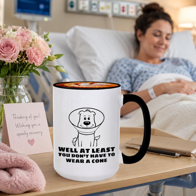 May include: A white ceramic mug with a black handle and rim, featuring a black line drawing of a dog wearing a cone collar. Below the illustration, the text reads: WELL AT LEAST YOU DON'T HAVE TO WEAR A CONE. The mug is filled with a brown beverage.