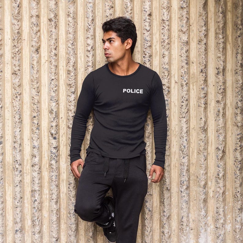 以下が含まれることがあります： 白い文字で「POLICE」と書かれた黒い長袖シャツ。シャツは黒いスウェットパンツと組み合わせています。人物は、質感のある明るい茶色の壁に寄りかかっています。