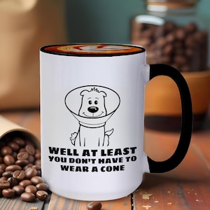 May include: White ceramic coffee mug with a black handle. The mug has a cartoon illustration of a dog wearing a cone and the text "Well at least you don't have to wear a cone".