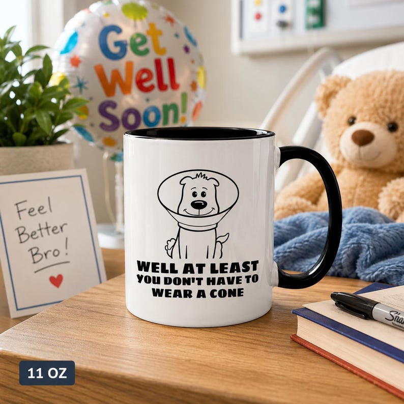 May include: A white ceramic mug with a black interior and handle, featuring a black line drawing of a dog wearing a cone collar and the text: WELL AT LEAST YOU DON'T HAVE TO WEAR A CONE. The mug has a capacity of 11 ounces as indicated by the text.