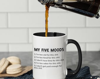 May include: White ceramic mug with a black handle and rim, featuring the text "MY FIVE MOODS" and a list of relatable sentiments. Coffee is being poured into the mug from a French press. The mug sits on a wooden coaster.