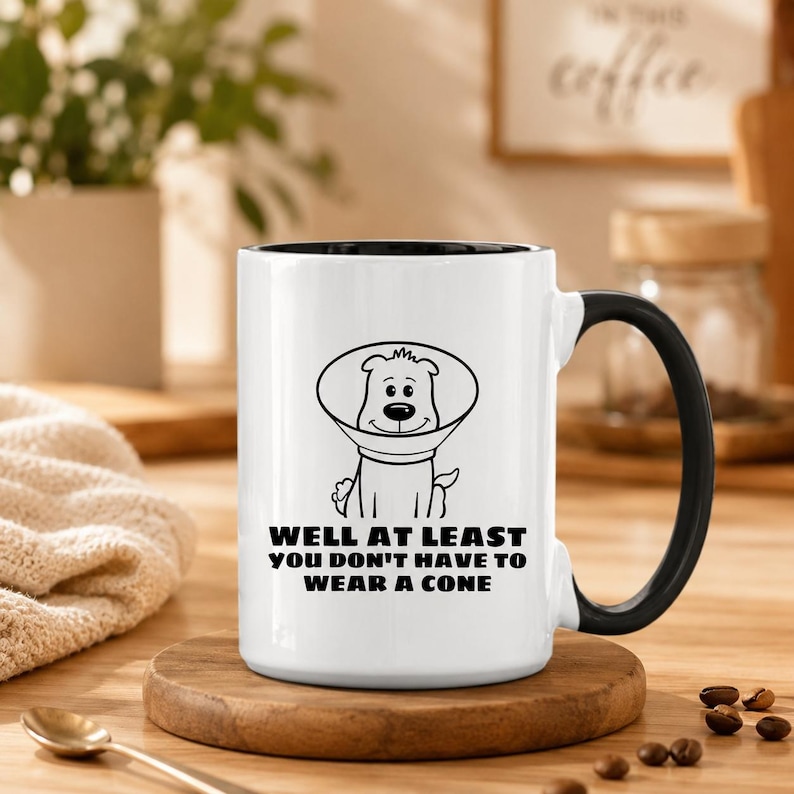 May include: A white ceramic mug with a black handle and interior. The front features a black line drawing of a dog wearing a veterinary cone, accompanied by the black text: WELL AT LEAST YOU DON'T HAVE TO WEAR A CONE printed below the illustration.