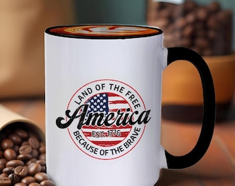 Könnte beinhalten: Weiße Keramik-Kaffeetasse mit schwarzem Henkel. Die Tasse hat ein rot-weiß-blaues amerikanisches Flaggen-Design mit dem Text "Land of the Free America Est. 1776 Because of the Brave".
