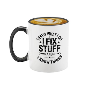 May include: White ceramic coffee mug with a black handle and rim, filled with coffee and latte art. The mug features the text "THAT'S WHAT I DO I FIX STUFF AND I KNOW THINGS" in black, with wrench and star graphics.