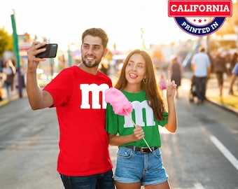 May include: A red t-shirt with a white letter m and a green t-shirt with a white letter m. The red shirt is worn with dark blue jeans, and the green shirt is worn with blue denim shorts. A circular badge reads Designed and Printed in California.