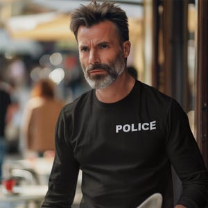 以下が含まれることがあります： 胸に白字で「POLICE」とプリントされた黒の長袖シャツ。グレーのひげと短いヘアスタイルをした人が着用しています。背景はぼやけており、屋外での設定を示唆しています。
