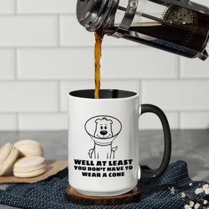 May include: A white ceramic mug with a black interior and handle, featuring a black line drawing of a dog wearing a cone and the text "WELL AT LEAST YOU DON'T HAVE TO WEAR A CONE" printed in black on the front of the mug.