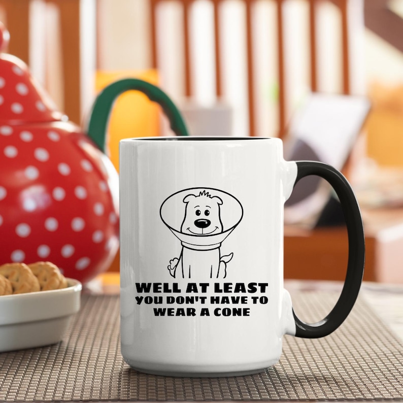 May include: A white ceramic mug with a black handle and interior. The front features a black line drawing of a dog wearing a veterinary cone, accompanied by the black text: WELL AT LEAST YOU DON'T HAVE TO WEAR A CONE printed in a bold sans-serif font.
