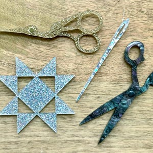 May include: A collection of craft items: gold glitter scissors, a silver needle, teal scissors, and a silver glitter star. The items are arranged on a wooden surface. The scissors are approximately 10 cm long.