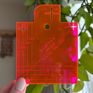 Puede incluir: Una guía de costura de plástico naranja brillante con un patrón de cuadrícula y marcas en pulgadas. La guía tiene un agujero en el centro y el texto "kimtsu seam guide kimtsu nit zin: comes in handy" impreso en ella.