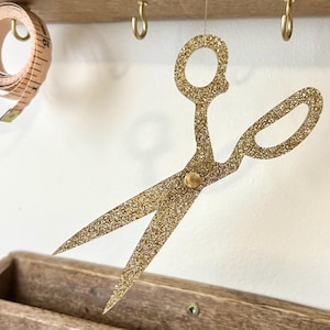 May include: A gold glitter scissors decoration hanging from a wooden shelf. A measuring tape is coiled on the left. The scissors are a craft or sewing-themed item, with a decorative design.