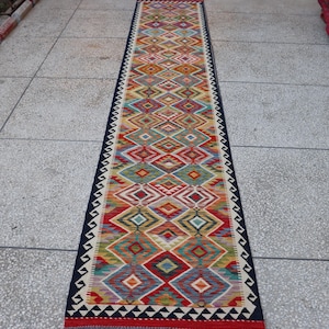 May include: A long, rectangular runner rug with a geometric diamond pattern in various colors, including red, yellow, blue, and green. The rug has a dark blue border with a zigzag design and fringed ends. The rug is likely made of woven fabric.