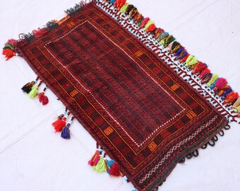 May include: A rectangular, hand-woven rug in shades of burgundy and brown, featuring a central striped pattern and a decorative border. The rug is adorned with colorful tassels along the edges, adding a vibrant touch to the design. The rug is approximately 24 inches long and 16 inches wide.