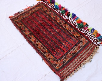 May include: A rectangular, hand-woven rug with a red and black geometric pattern. The rug has a border with tan and brown details and colorful tassels along one edge. The rug is approximately 24 inches long and 16 inches wide.